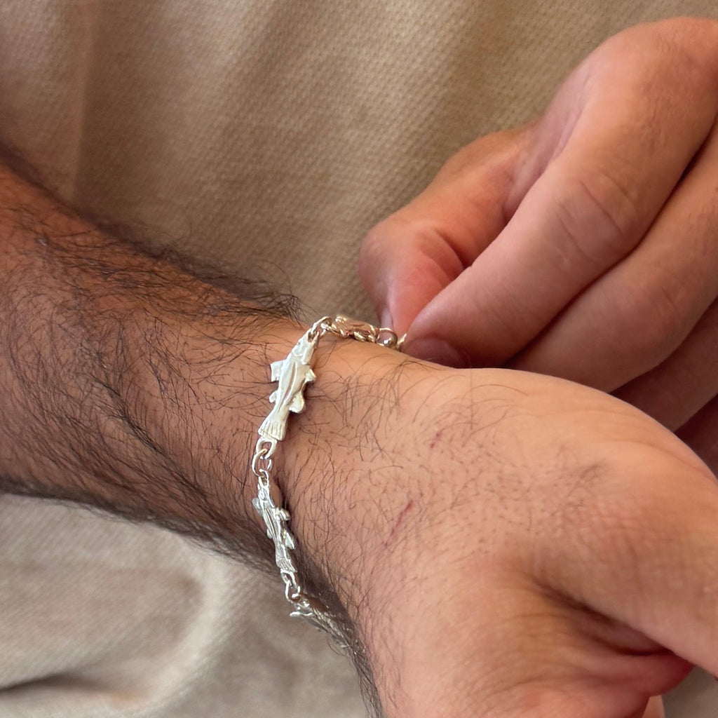 A close-up of a person's wrist wearing a delicate silver bracelet with a fish design. The person is wearing a beige t-shirt.